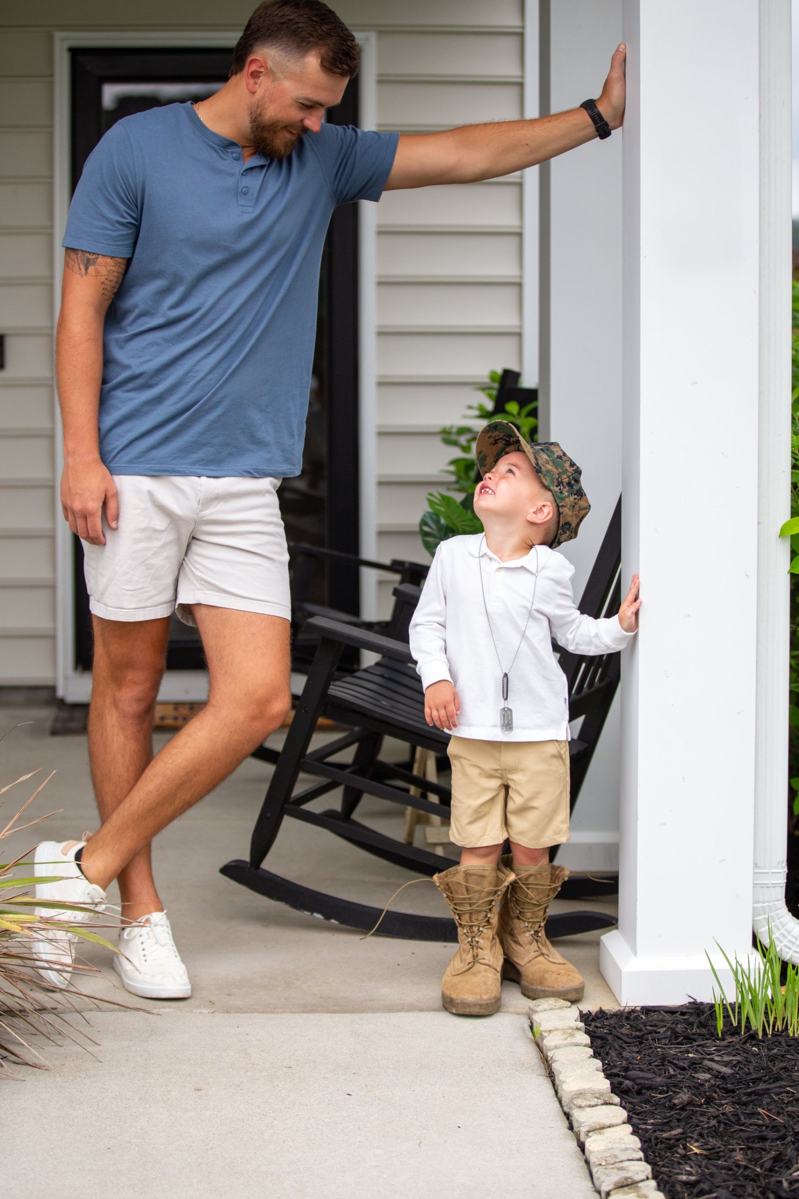 A Marine veteran smiles at his young son wearing his cover and boots on the front porch