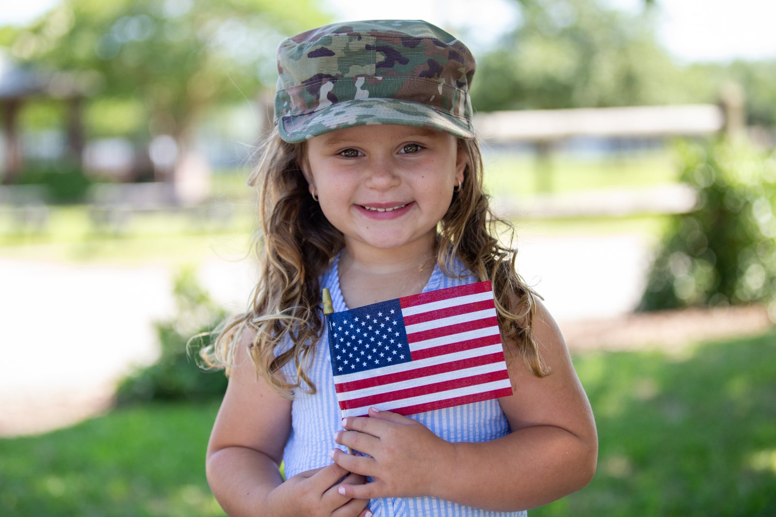 A smiling military child wearing a camouflage cap holds a small American flag